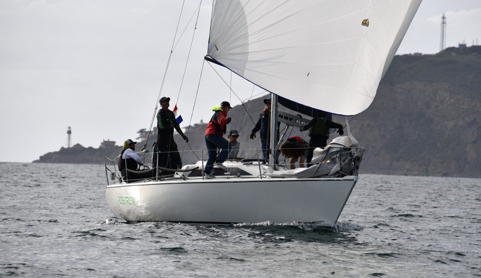 Racing yacht Positron sailing under full canvas past a coastal lighthouse